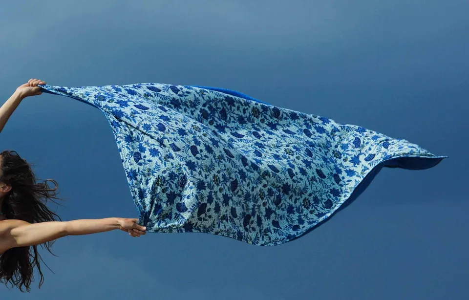 Femme bras levés déployant un grand foulard imprimé bleu dans le vent devant une mer sombre, à Porquerolles.