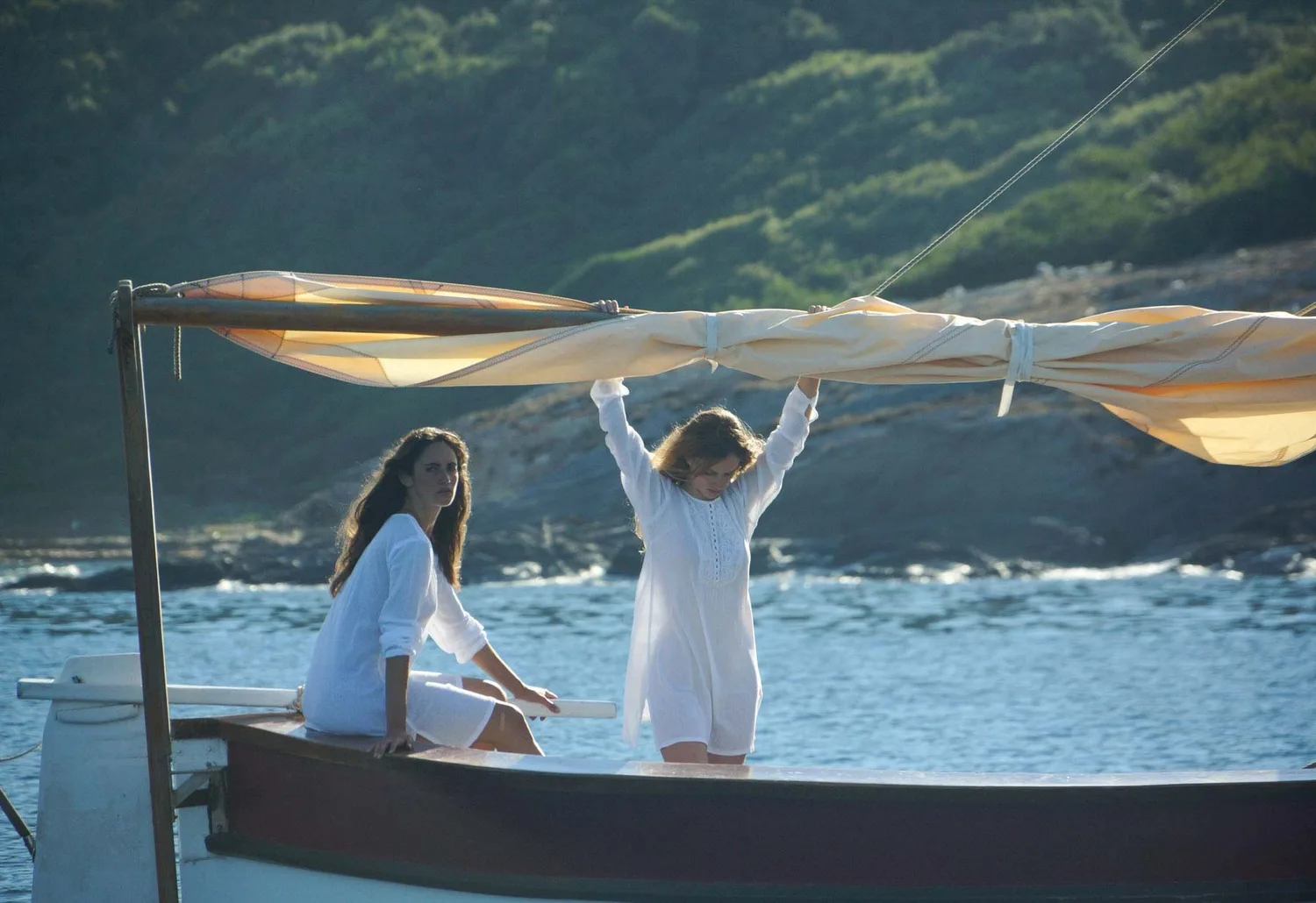 Deux femmes en coton blanc sur un pointu, l'une debout bras levés sous la voile, côte verte au fond, à Porquerolles.
