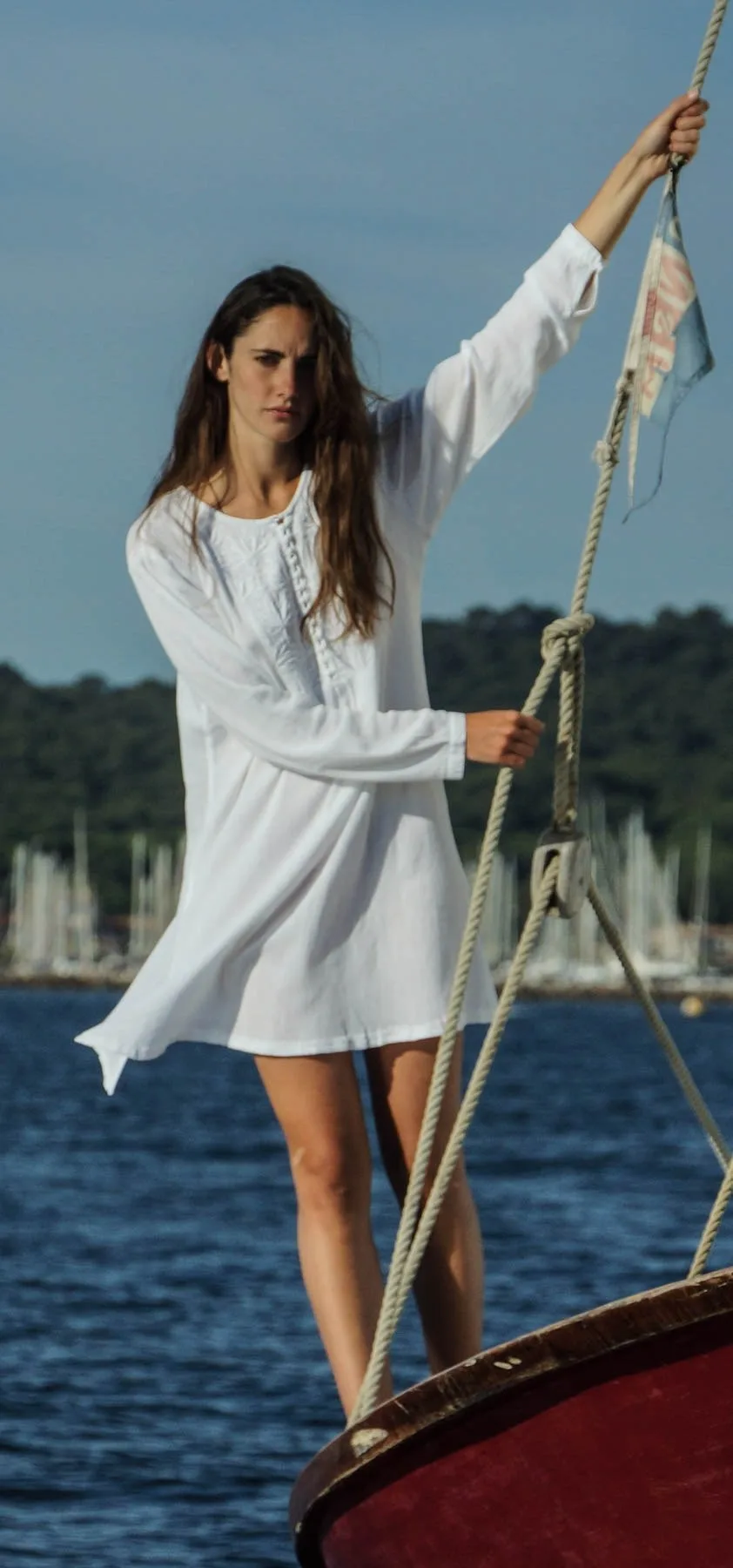 Femme en tunique de coton blanc debout à la proue d'un bateau, main au cordage, mâts du port derrière, à Porquerolles.