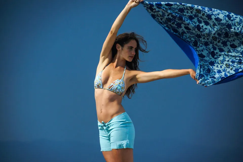 Femme en bikini fleuri tenant un foulard bleu à motifs qui claque dans le vent, mer sombre en fond, à Porquerolles.