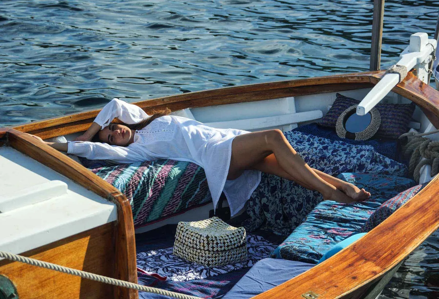 Femme allongée en tunique de coton blanc brodé sur les coussins d'un bateau, sac en vannerie sur le pont, à Porquerolles.
