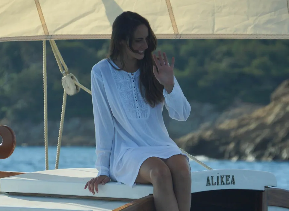 Femme en tunique de coton blanc brodé debout à la proue près du mât, main levée, nom du bateau visible, à Porquerolles.
