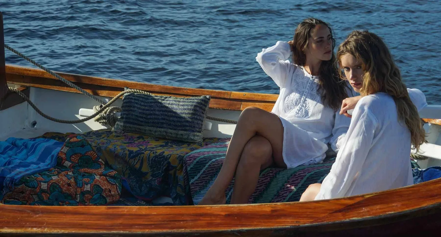 Deux femmes en tuniques de coton blanc assises sur les coussins colorés d'un bateau, la mer en fond, à Porquerolles.