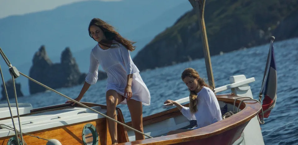 Deux femmes en coton blanc sur un bateau, l'une descendant du plat-bord, l'autre assise, rochers de la côte au fond, à Porquerolles.