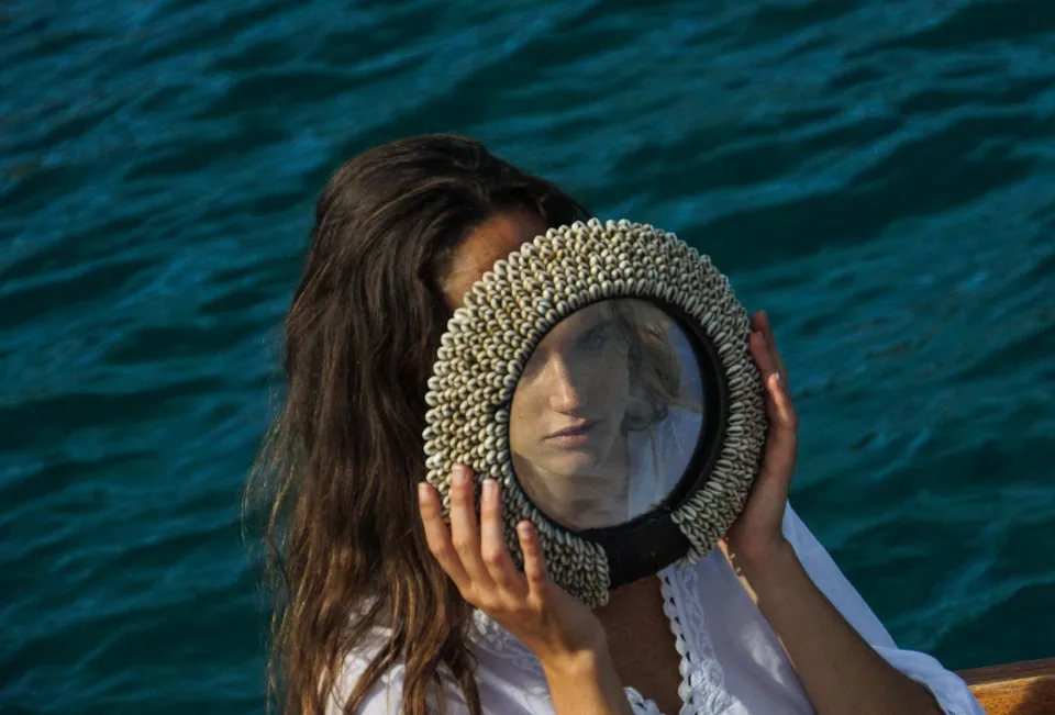 Femme en chemise de coton et dentelle tenant un miroir cerclé de coquillages devant son visage, mer sombre en fond, à Porquerolles.