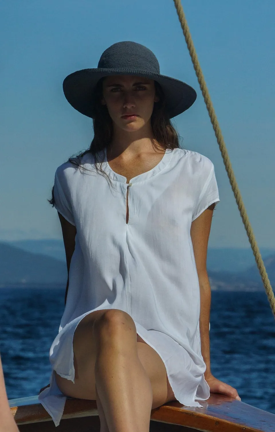 Femme en chapeau de paille gris et chemise de coton blanc assise à la proue, regard face caméra, côte dans la brume, à Porquerolles.