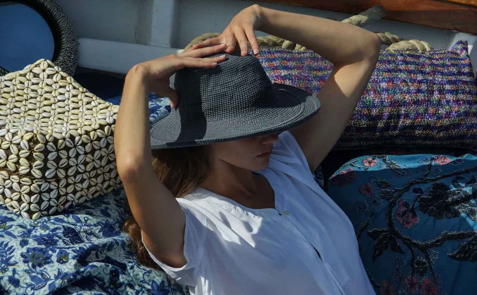 Femme en chemise de coton blanc allongée sur les coussins, les deux mains ajustant son chapeau de paille gris, à Porquerolles.