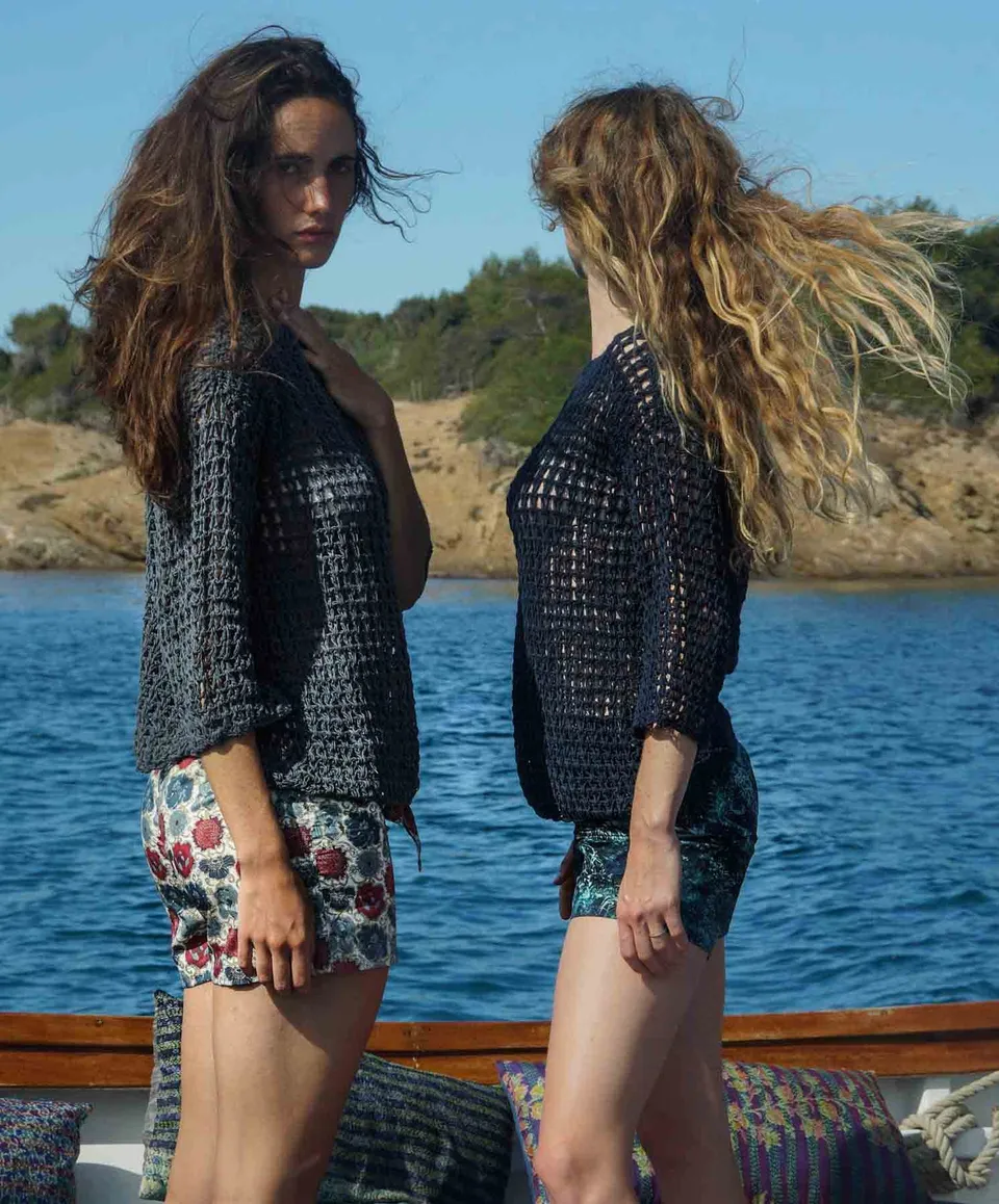 Deux femmes face à face en pulls de crochet marine et shorts imprimés, bastingage du bateau, côte rocheuse en fond, à Porquerolles.