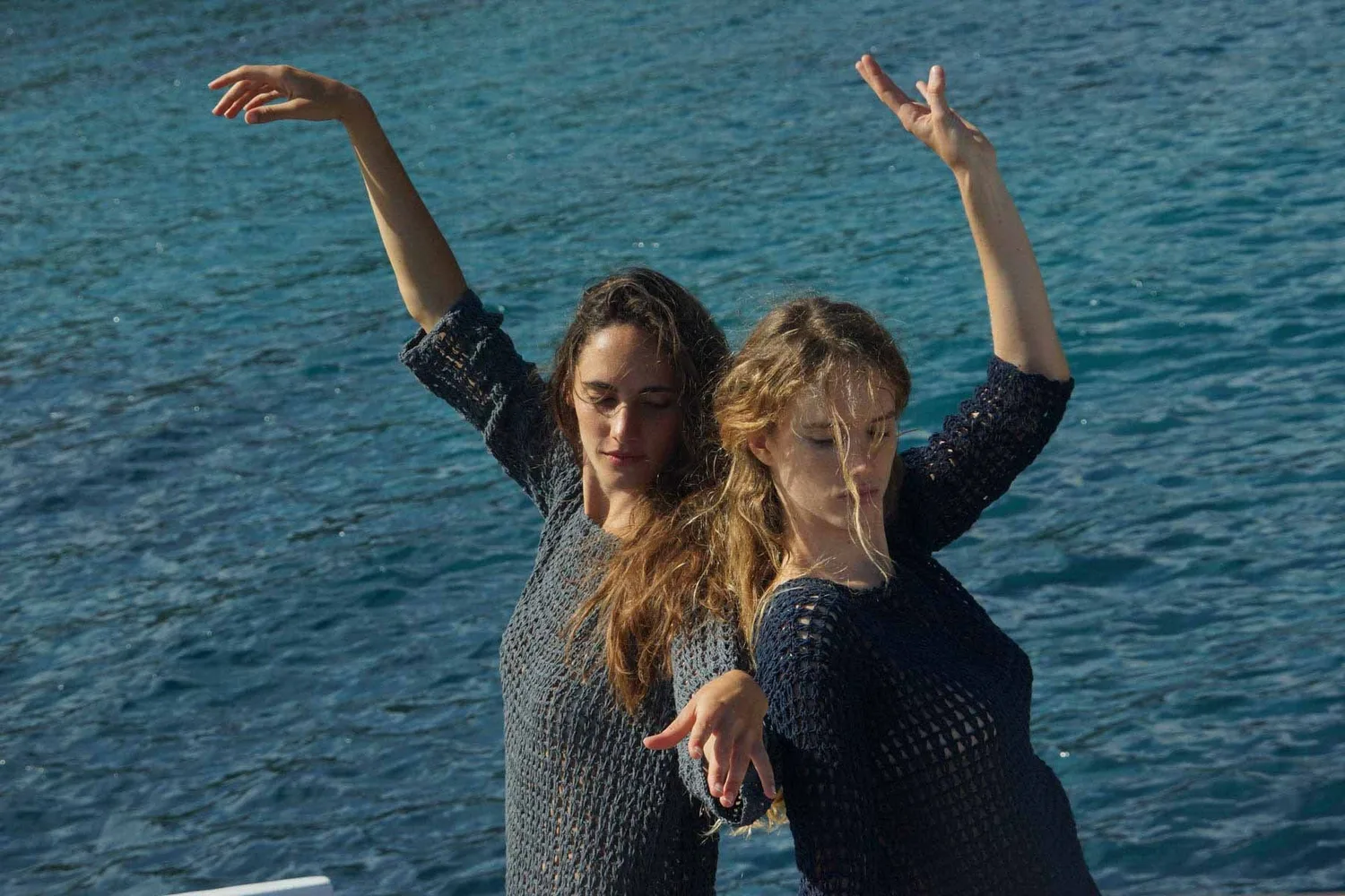 Deux femmes en pulls de crochet marine, bras levés dos à dos dans un mouvement de danse, mer en fond, à Porquerolles.