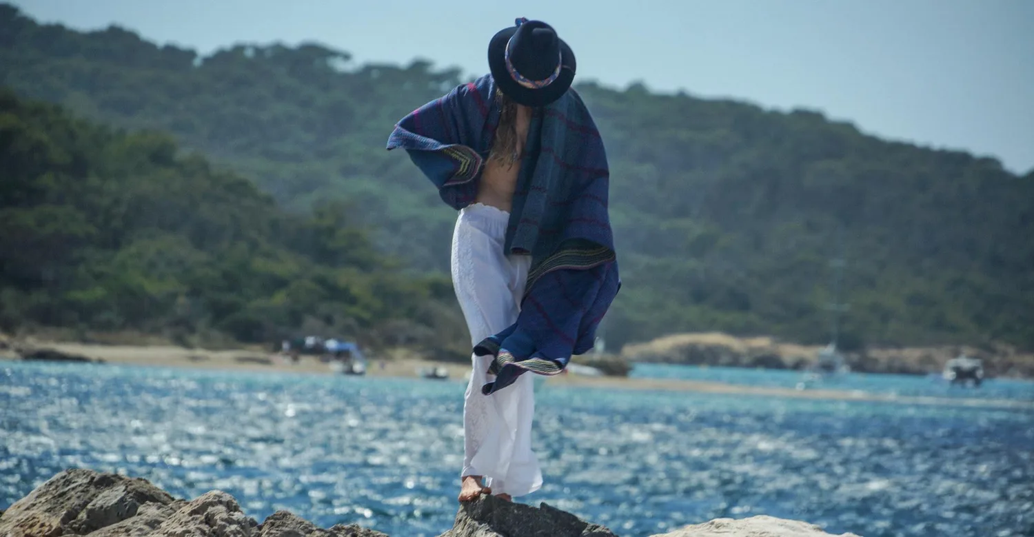 Femme sur les rochers en pantalon de coton blanc brodé, chapeau noir et poncho tissé bleu couvrant le visage, plage et bateaux en fond, à Porquerolles.