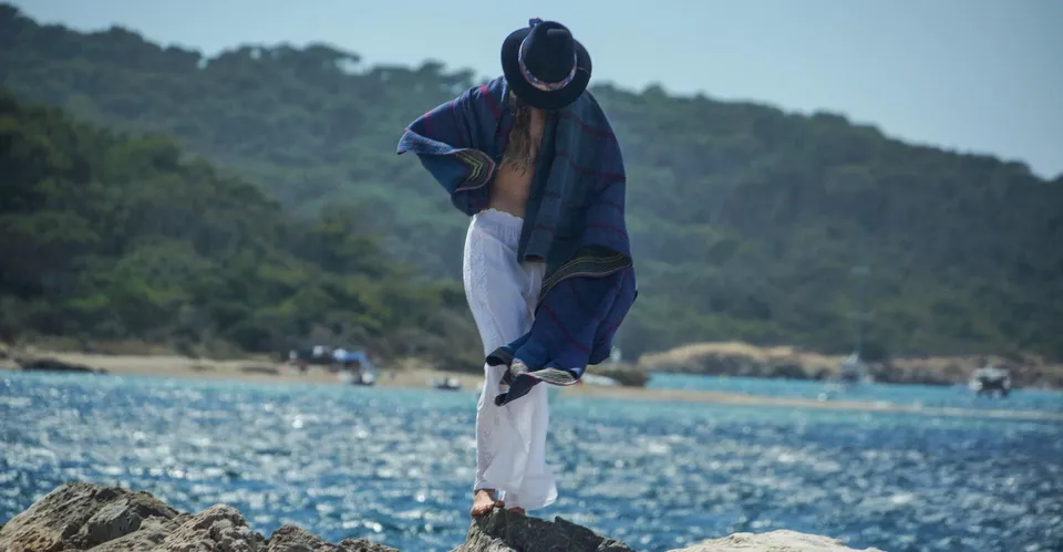 Femme sur les rochers en pantalon de coton blanc brodé, chapeau noir et poncho tissé bleu couvrant le visage, plage et bateaux en fond, à Porquerolles.
