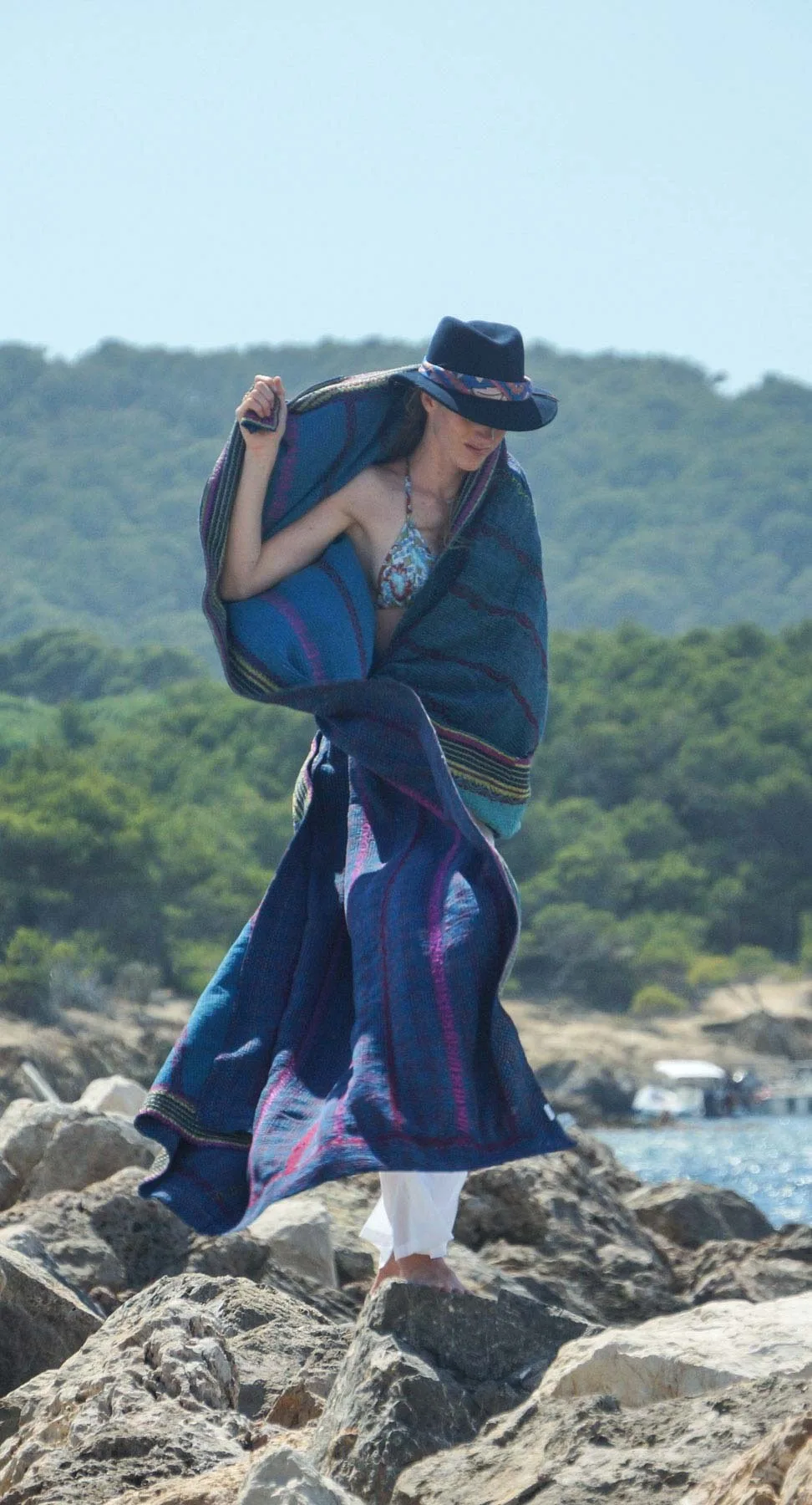 Femme sur les rochers en bikini et chapeau noir, un poncho tissé bleu drapé sur les épaules, à Porquerolles.