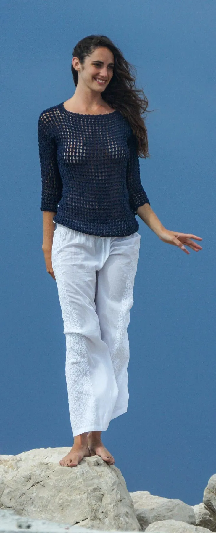 Femme en pull de crochet marine et pantalon de coton blanc, bras levés sur la digue, ciel d'orage spectaculaire en fond, à Porquerolles.