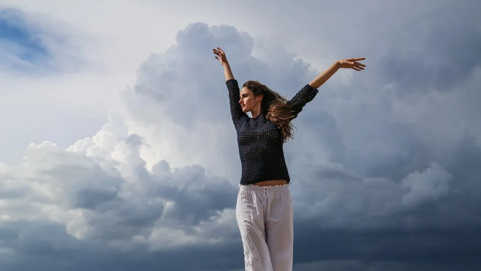 Femme en pull de crochet marine et pantalon de coton blanc, bras levés sur la digue, nuages d'orage menaçants, à Porquerolles.