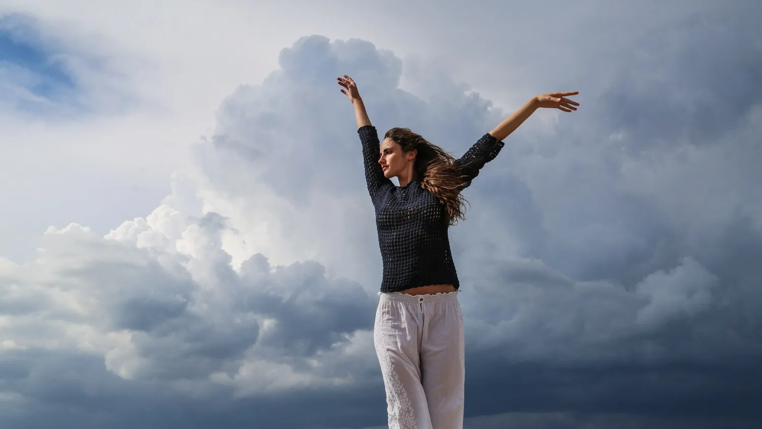Femme en pull de crochet marine et pantalon de coton blanc, bras levés sur la digue, nuages d'orage menaçants, à Porquerolles.