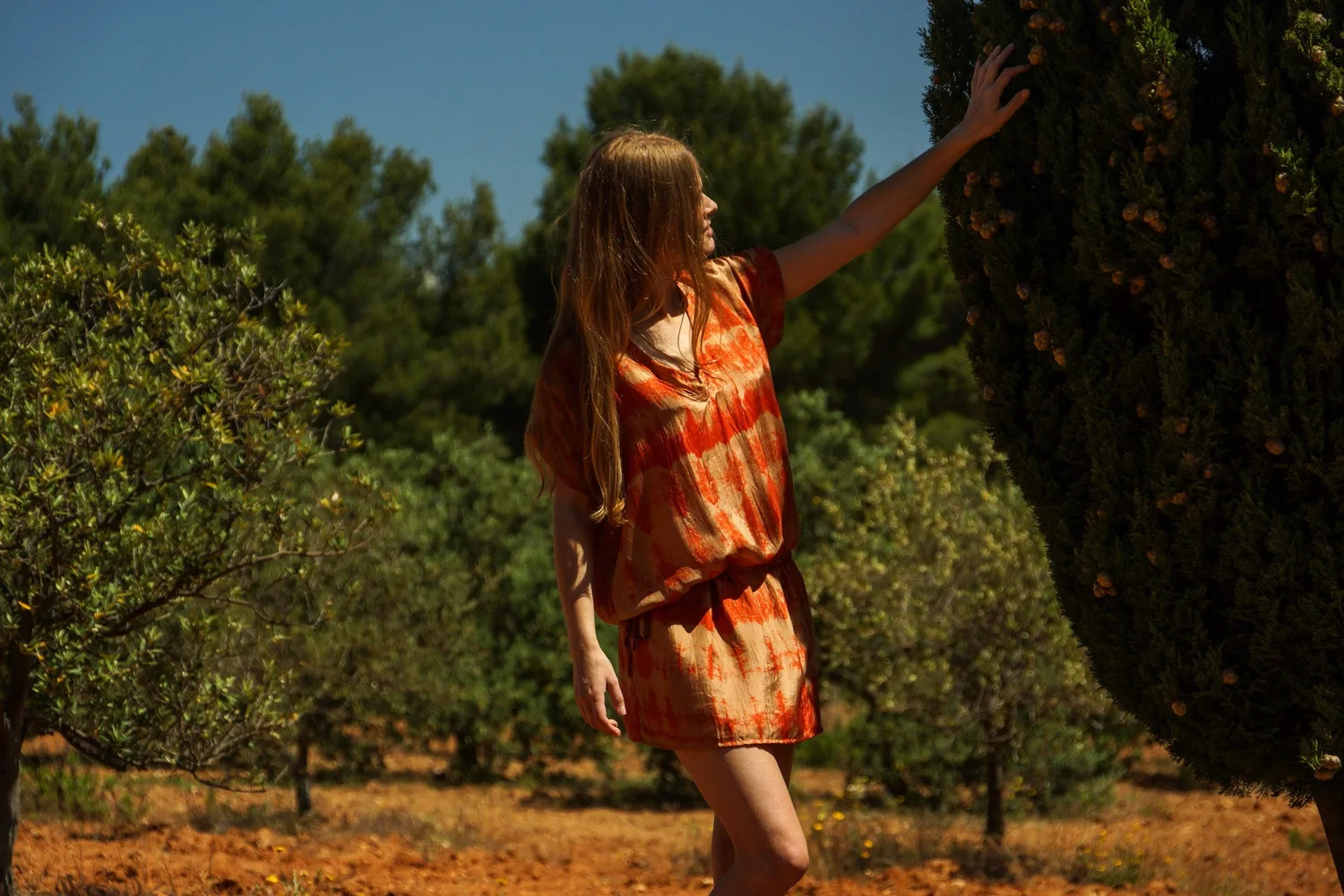 Femme de profil en tunique tie-dye orange touchant un cyprès dans une oliveraie ensoleillée à Porquerolles.