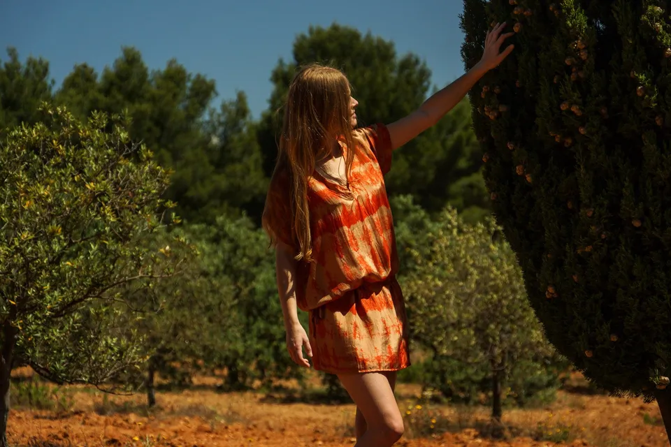 Femme de profil en tunique tie-dye orange touchant un cyprès dans une oliveraie ensoleillée à Porquerolles.