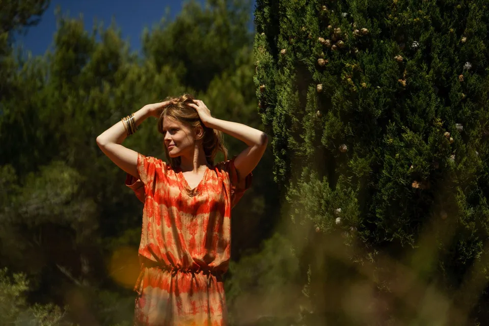 Femme en tunique orange à motifs géométriques, mains dans les cheveux, maquis de Porquerolles derrière.