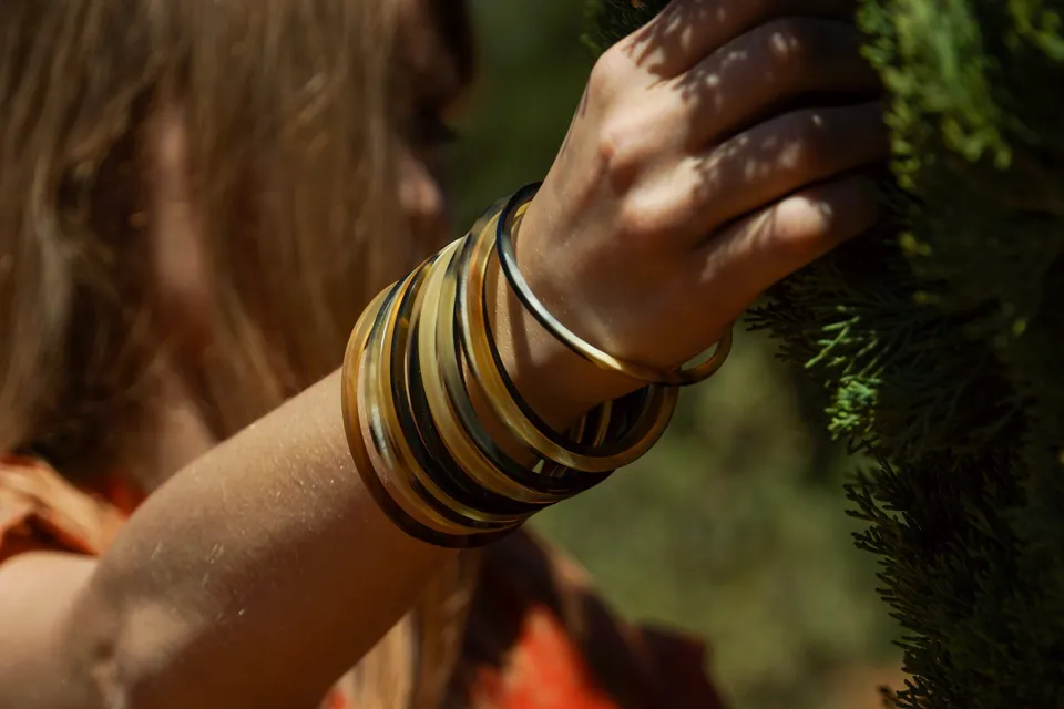 Détail de bracelets fins de laiton au poignet, main posée sur un feuillage vert.