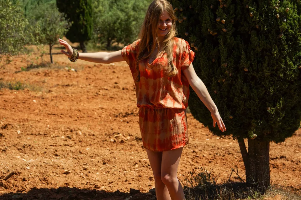 Femme en tunique orange tie-dye, bras écartés sur terre ocre devant le maquis et les oliviers à Porquerolles.