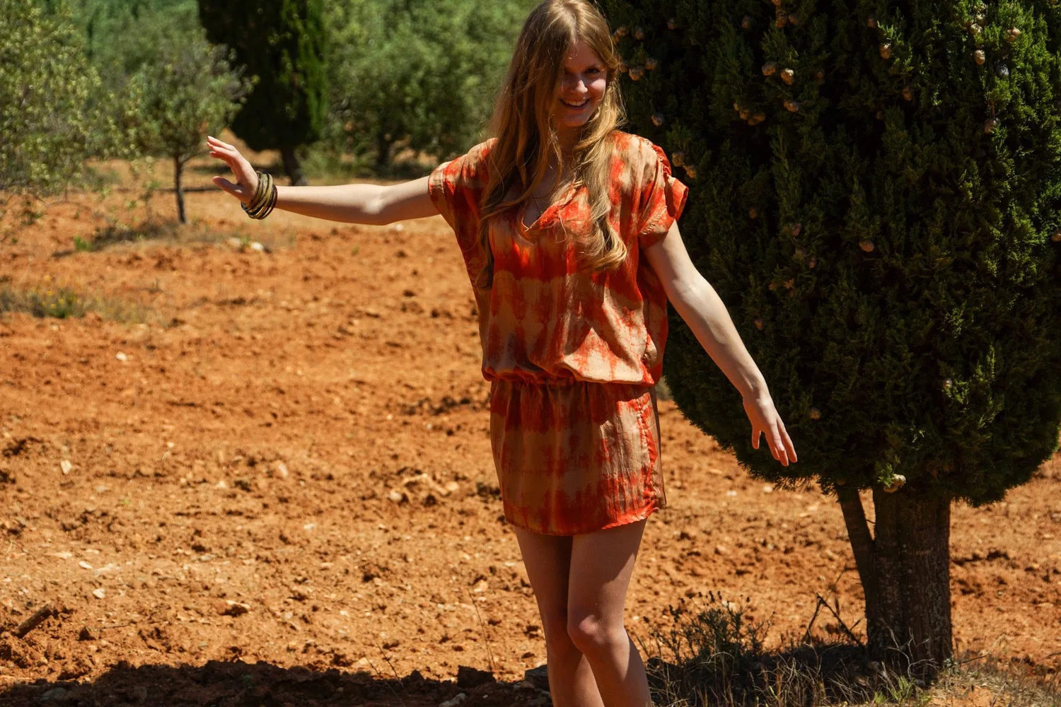 Femme en tunique orange tie-dye, bras écartés sur terre ocre devant le maquis et les oliviers à Porquerolles.