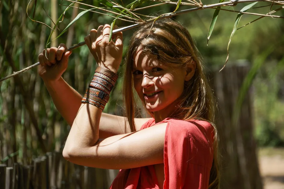 Femme souriante en tunique corail et bracelet tressé, main sur une branche du maquis ensoleillé à Porquerolles.