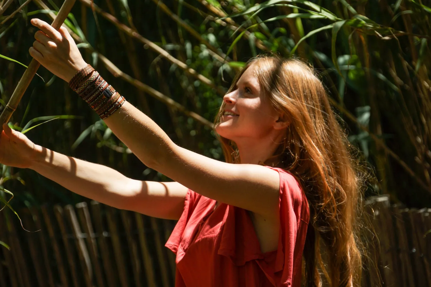 Femme souriante en robe corail, bras tendus vers une barrière de roseaux dans un jardin.