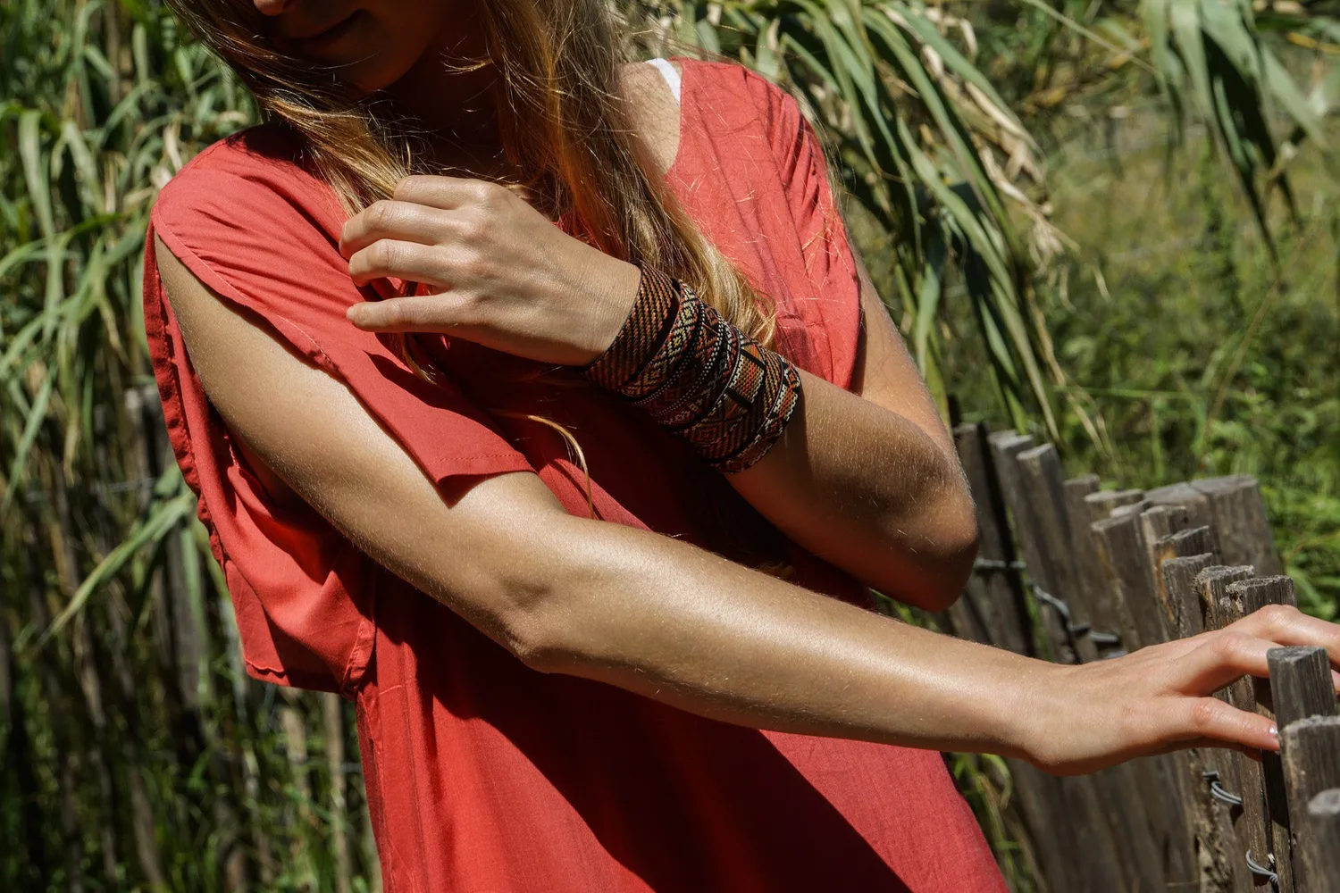 Femme en tunique terracotta et bracelet tissé appuyée à une clôture de bois parmi les roseaux à Porquerolles.