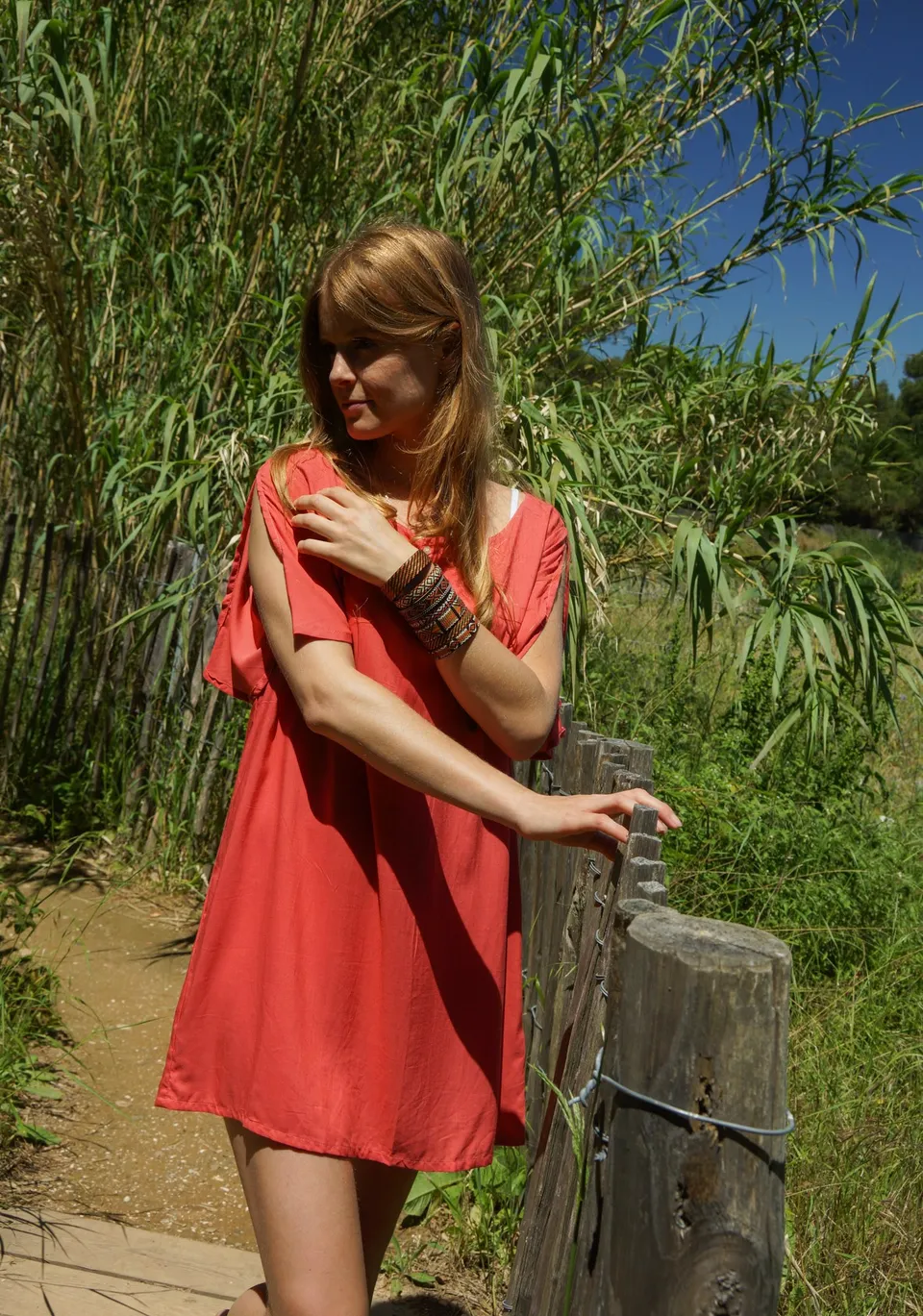Femme en robe corail appuyée à un poteau de bois le long d'une barrière de roseaux.