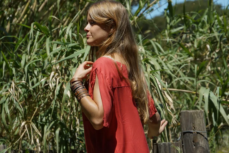 Femme de profil en tunique corail et bracelets tissés près de roseaux hauts sous un ciel bleu à Porquerolles.