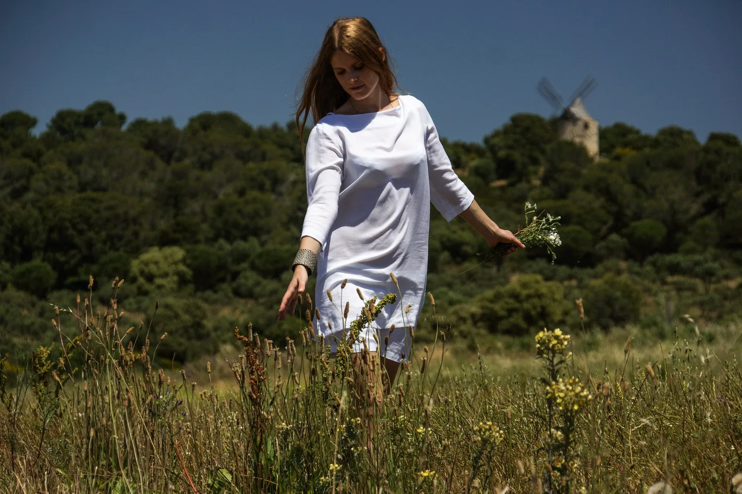 Femme en tunique de lin blanc effleurant les hautes herbes d'un champ de marguerites.