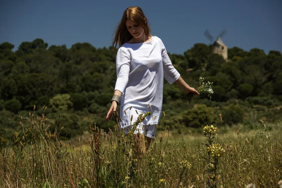 Femme en tunique de lin blanc effleurant les hautes herbes d'un champ de marguerites.