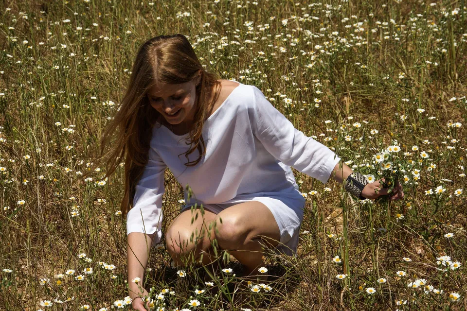 Femme en tunique de lin blanc accroupie dans les marguerites, cueillant un bouquet.
