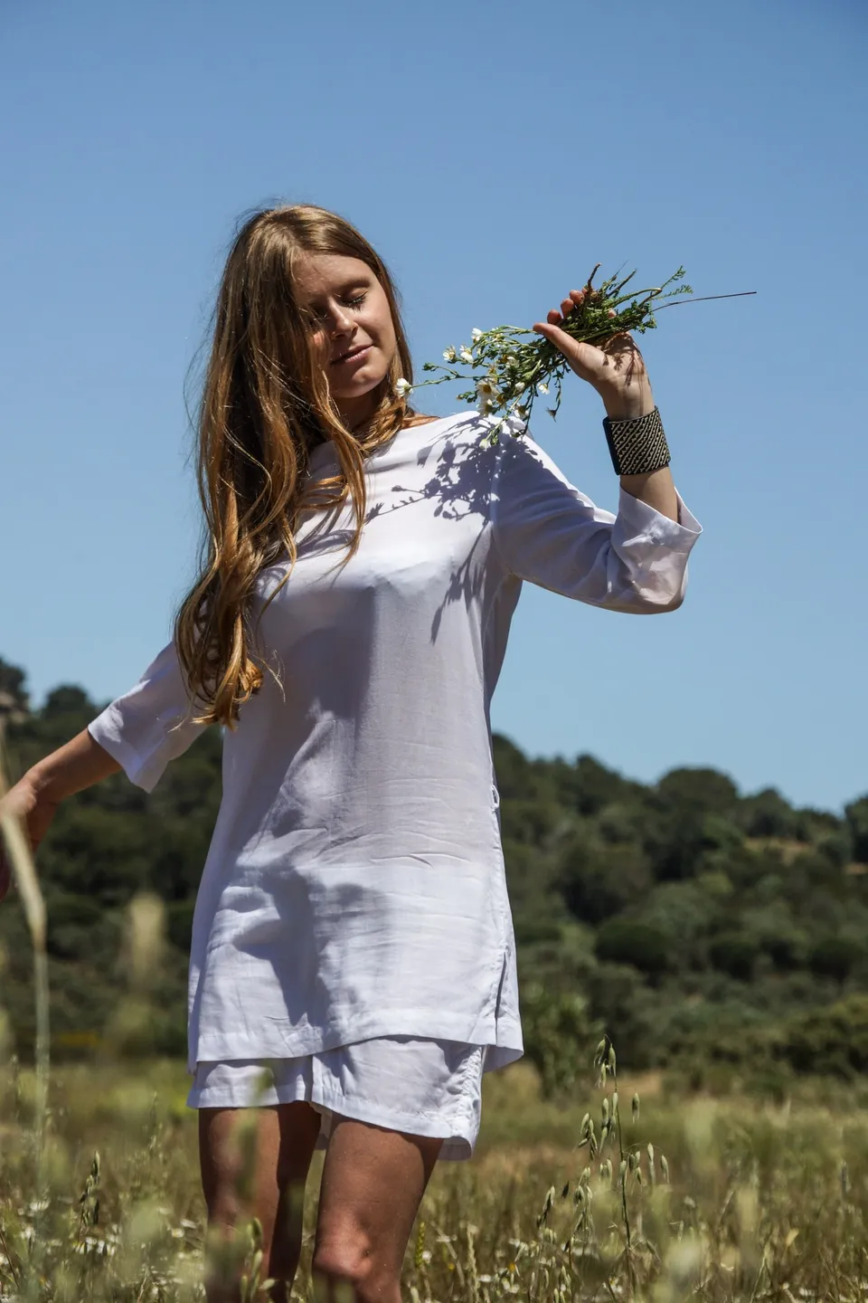 Femme en tunique de lin blanc tenant un bouquet de fleurs sauvages dans un champ herbacé sous ciel bleu à Porquerolles.