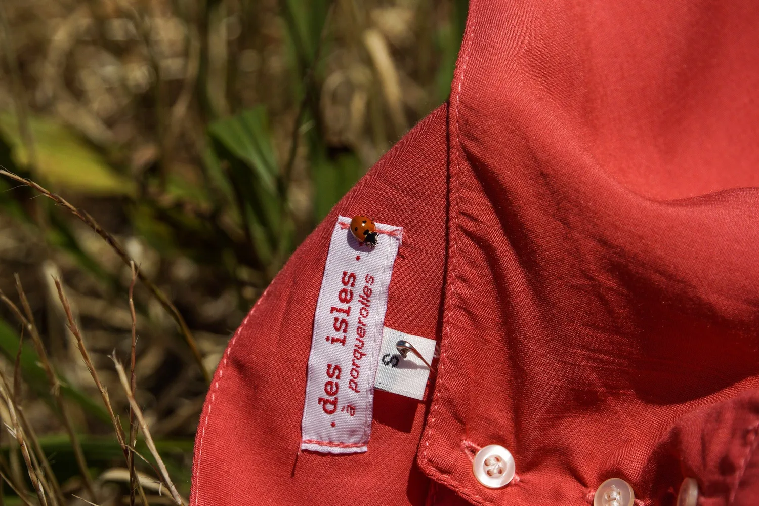 Détail de l'étiquette « des isles · porquerolles » cousue au col d'une chemise corail, une coccinelle posée dessus.