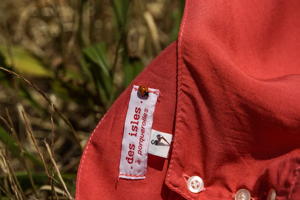 Détail de l'étiquette « des isles · porquerolles » cousue au col d'une chemise corail, une coccinelle posée dessus.