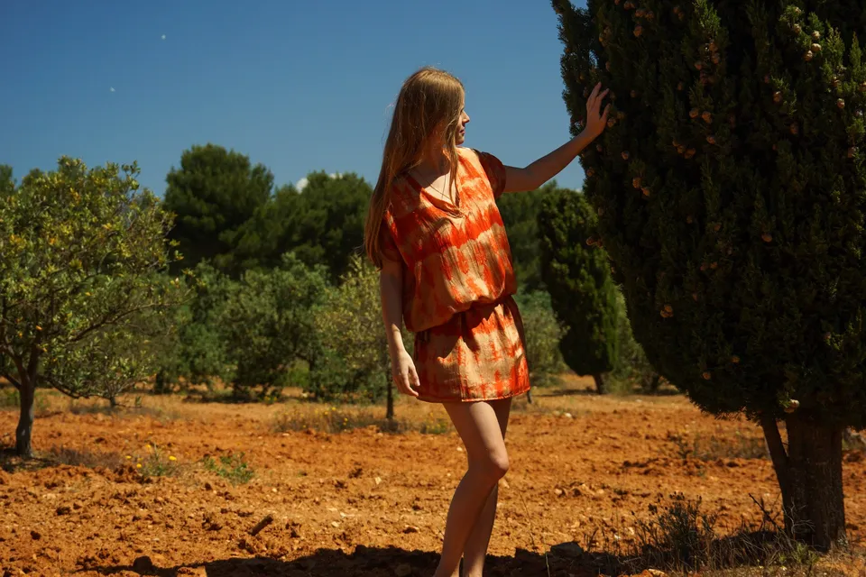 Femme en tie-dye orange, main tendue vers un cyprès, terre rouge du maquis de Porquerolles.