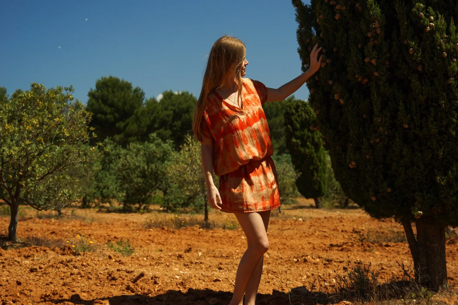 Femme en tie-dye orange, main tendue vers un cyprès, terre rouge du maquis de Porquerolles.